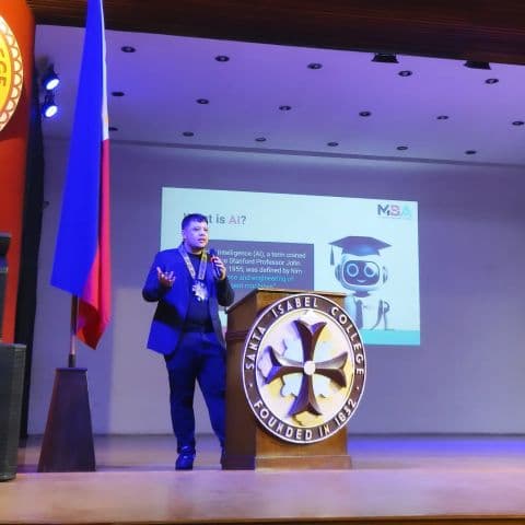 JC delivering a keynote on AI at Santa Isabel College, speaking from the podium with microphone
